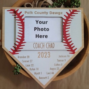 May include: A baseball-themed photo frame in the shape of home plate. The frame is light wood with a white and red baseball stitch design. Text includes "Polk County Dawgz", "Your Photo Here", "COACH CHAD 2023", and player names with numbers.
