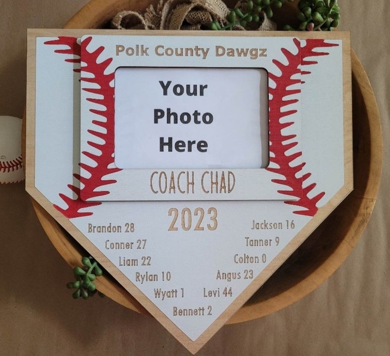 May include: A baseball-themed photo frame in the shape of home plate. The frame is light wood with a white and red baseball stitch design. Text includes "Polk County Dawgz", "Your Photo Here", "COACH CHAD 2023", and player names with numbers.