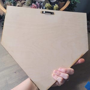 May include: A light-colored, unfinished wooden plaque in a baseball home plate shape. The back of the plaque has a metal sawtooth hanger. The plaque is held by a hand, with a blurred background of a wooden bowl with plants.
