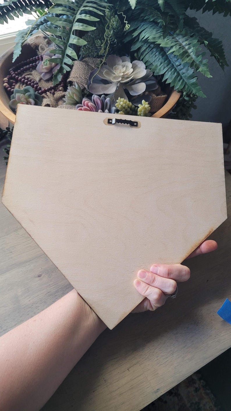 May include: A light-colored, unfinished wooden plaque in a baseball home plate shape. The back of the plaque has a metal sawtooth hanger. The plaque is held by a hand, with a blurred background of a wooden bowl with plants.