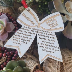 May include: A wooden bow with the text "Destinee" and "MHS Class of 2025" on it. The bow has a message about hard work, dedication, and victories.