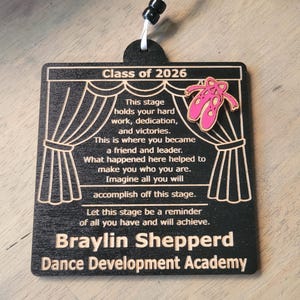May include: A black wooden square ornament featuring a stage curtain design, pink ballet slippers, and the text: Class of 2026, This stage holds your hard work, dedication, and victories. This is where you became a friend and leader. Braylin Shepperd Dance Development Academy.