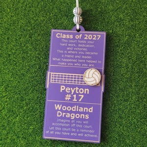 May include: A rectangular purple commemorative plaque featuring a white volleyball graphic, a net, and text including Class of 2027, Peyton #17, and Woodland Dragons. The plaque hangs from a white ribbon with two small spherical beads featuring volleyball patterns.