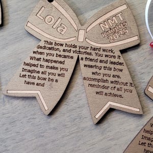 May include: A wooden bow tie ornament with the text "Lola" on one side and "NMT Knights 2025" on the other side. The ornament has a message about hard work, dedication, and victories.