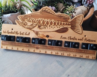 Könnte beinhalten: Wanddekoration aus Holz in Fischform mit einem Lineal und Haken zum Aufhängen von Angelködern. Das Lineal ist mit dem Text "Bait", "Throw Back", "Keeper", "Awesome Story" und "A Lie" beschriftet. Der Text "We hooked the best dad!" und "Love, Charles and Harrison" ist ebenfalls in das Holz graviert.