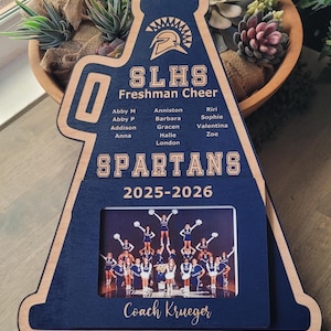 May include: A navy blue and natural wood megaphone-shaped plaque featuring the text SLHS Freshman Cheer, the word SPARTANS, the year 2025-2026, and Coach Krueger. It includes a list of names and a rectangular photo frame displaying a cheerleading team.