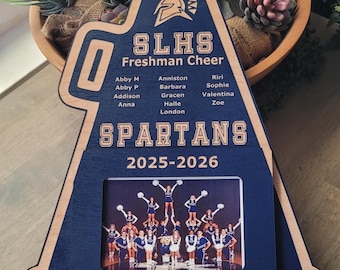 May include: A navy blue and natural wood megaphone-shaped plaque featuring the text SLHS Freshman Cheer, the word SPARTANS, the year 2025-2026, and Coach Krueger. It includes a list of names and a rectangular photo frame displaying a cheerleading team.