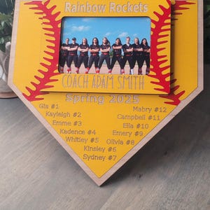 May include: A wooden softball-shaped plaque with a photo of a team, "Rainbow Rockets," and the text "Coach Adam Smith Spring 2025." The plaque is yellow with red stitching and includes player names and numbers.