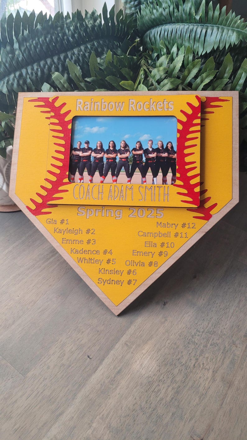 May include: A wooden softball-shaped plaque with a photo of a team, "Rainbow Rockets," and the text "Coach Adam Smith Spring 2025." The plaque is yellow with red stitching and includes player names and numbers.