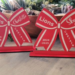 May include: Two red wooden cheer bow cutouts with white accents. The bows are decorated with the text "Lions", "Captain", "2024-2025", "Charlotte" and "Skye".