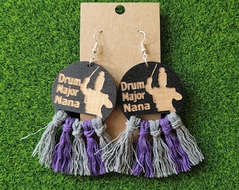 May include: A pair of circular black wooden earrings featuring the engraved text Drum Major Nana and a silhouette of a drum major. Each earring is accented with a multi-strand tassel fringe in alternating shades of grey and purple yarn.