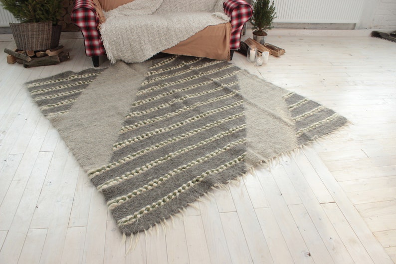May include: A gray and white striped wool rug with a geometric pattern. The rug has a fringed edge and is laid out on a white wood floor.