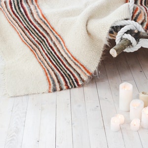 May include: A white blanket with a colorful striped pattern is draped over a wooden branch. White candles are scattered on a white wooden floor.