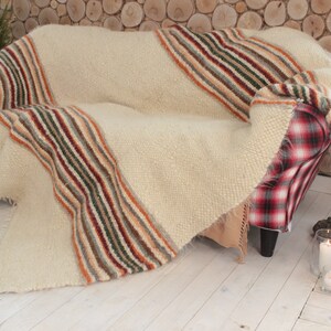 May include: A white throw blanket with multi-colored stripes draped over a red and black plaid armchair. The blanket has a fluffy texture and is made of wool.