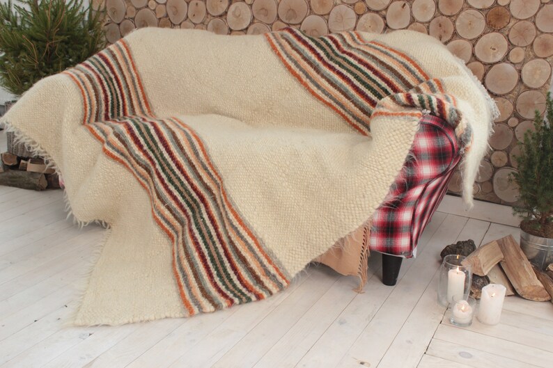 May include: A white throw blanket with multi-colored stripes draped over a red and black plaid armchair. The blanket has a fluffy texture and is made of wool.