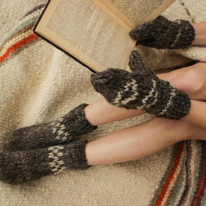May include: A person wearing brown and white knitted mittens and socks while reading a book on a white and orange striped blanket.