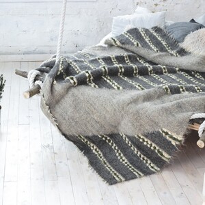May include: A gray and white striped blanket draped over a wooden bed frame with white rope hanging from the ceiling. The bed is in a white room with a small potted tree and candles on the floor.