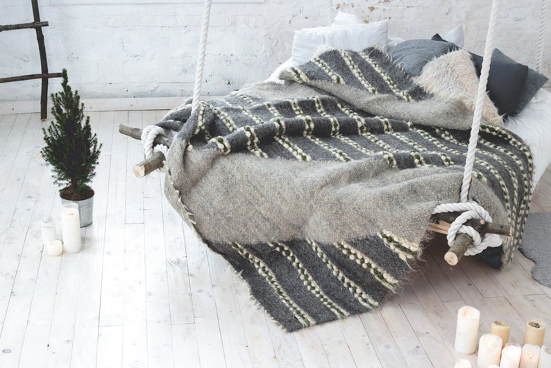 May include: A gray and white striped blanket draped over a wooden bed frame with white rope hanging from the ceiling. The bed is in a white room with a small potted tree and candles on the floor.