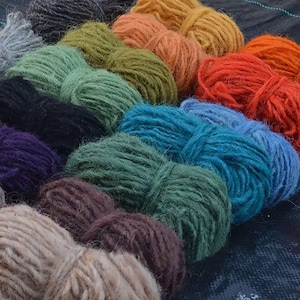 Op de afbeelding: Een verzameling garenklossen in verschillende kleuren, waaronder naturel, groen, blauw, paars, bruin en oranje. Het garen is in een rij gerangschikt, met de donkerste kleuren onderaan en de lichtste kleuren bovenaan.