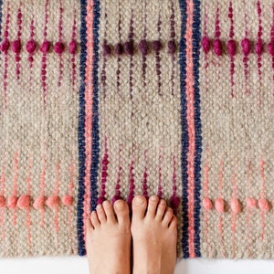 May include: A woven rug with a beige background and vertical stripes in pink and navy blue. The rug has decorative pink and purple bobbles. Bare feet are visible at the bottom of the rug.