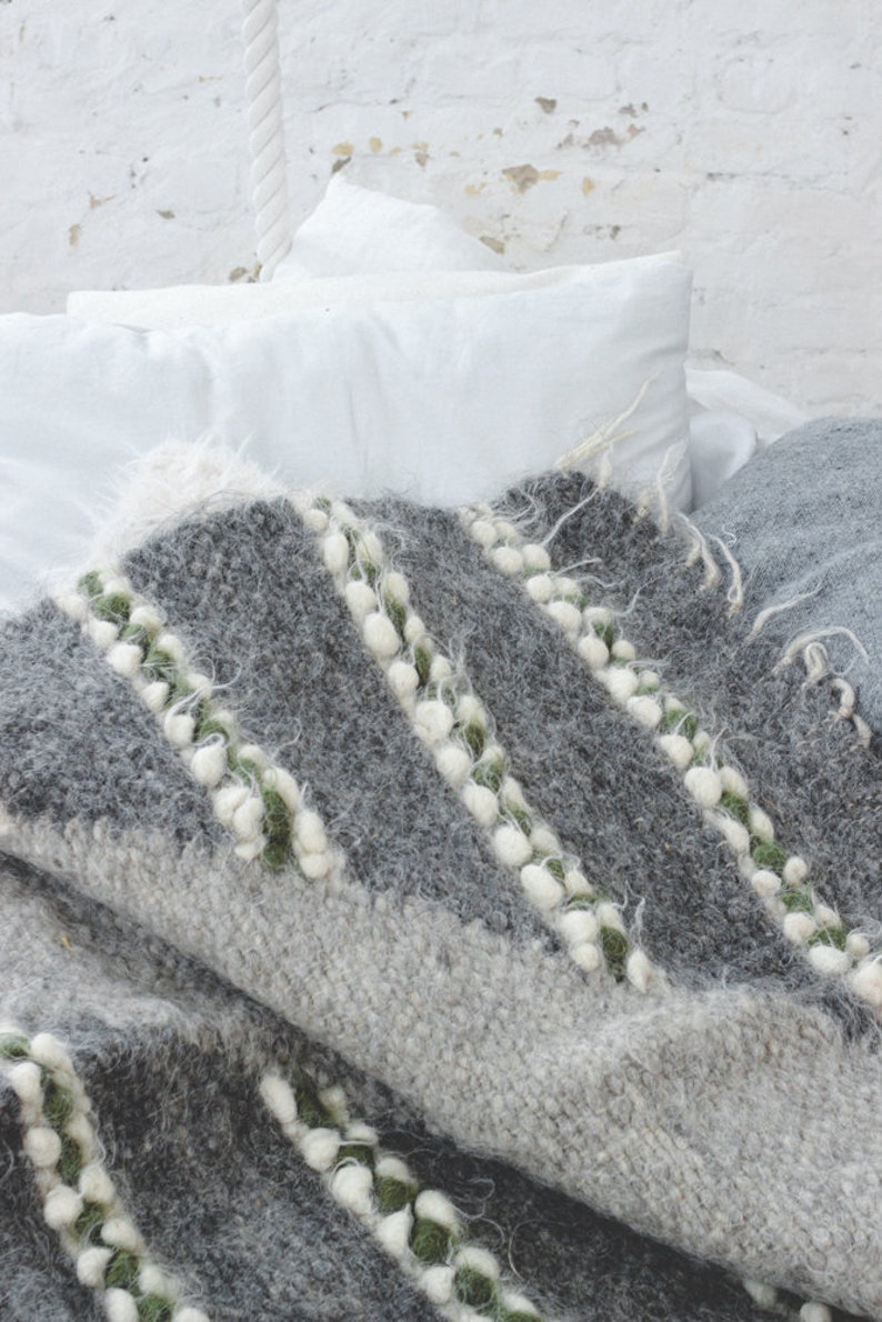 May include: A gray and white woven throw blanket with green accents. The blanket has a textured, fuzzy surface and is folded on a bed.