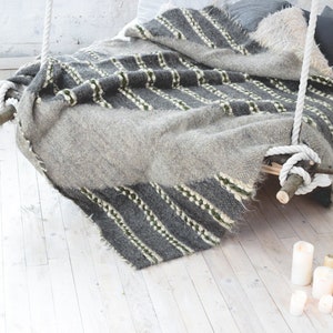 May include: A gray and white striped blanket with green accents is draped over a wooden bed frame with white rope hanging from the ceiling. The bed is sitting on a white wood floor with white candles scattered around it.