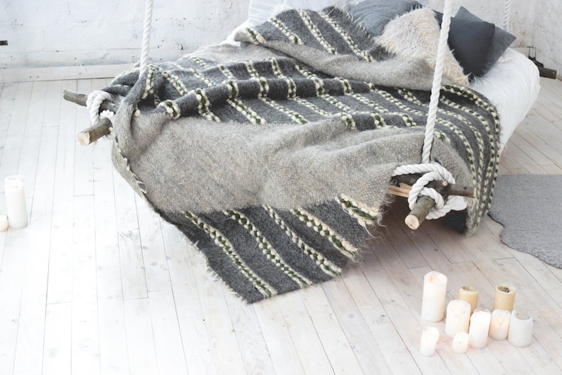 May include: A gray and white striped blanket with green accents is draped over a wooden bed frame with white rope hanging from the ceiling. The bed is sitting on a white wood floor with white candles scattered around it.