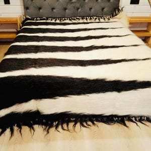 Puede incluir: Una manta grande a rayas blancas y negras cubre una cama. La manta tiene gruesas rayas horizontales y un fleco deshilachado. La cama está en una habitación con suelo de madera clara y paredes con paneles blancos. Dos pequeñas mesitas de noche de madera flanquean la cama.