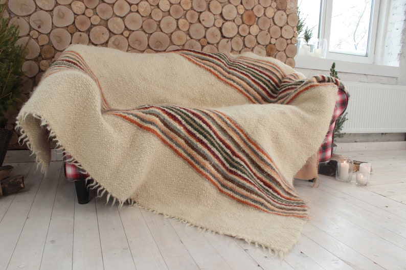 May include: A white and cream striped throw blanket with a fringe edge, draped over a couch with a red and white plaid pattern. The couch is in front of a wall made of stacked wooden rounds.