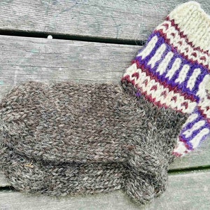 Puede incluir: Calcetines de lana tejidos a mano en tonos grises y blancos, con un puño estampado en morado, blanco y burdeos. Los calcetines son gruesos y parecen cálidos, perfectos para el frío. Están sobre una superficie de madera envejecida.
