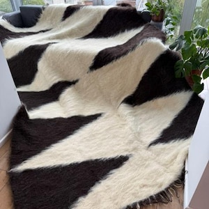 Puede incluir: Una manta rectangular grande con un patrón geométrico audaz. La manta presenta rayas alternas de color marrón oscuro y blanco roto, creando un contraste visual llamativo. El material parece ser lana suave y texturizada o una tela similar, con un borde con flecos.