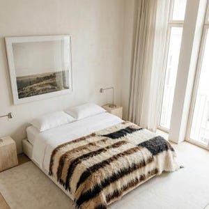 Pode incluir: Um quarto com uma cama coberta por um cobertor de pele sintética listrado em tons de marrom, preto e creme. O quarto tem uma paleta de cores neutras, com paredes brancas, um tapete de cor clara e uma impressão de paisagem emoldurada.