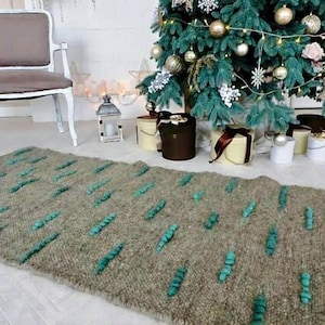 Puede incluir: Una alfombra tejida verde y marrón con una superficie texturizada. La alfombra es larga y rectangular y está decorada con pequeños pompones verdes.