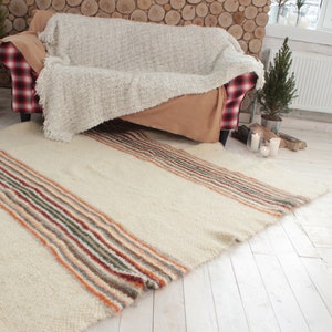 May include: A white, fluffy rug with colorful stripes in shades of orange, brown, green, and gray. The rug is laid out on a white wood floor in front of a couch.