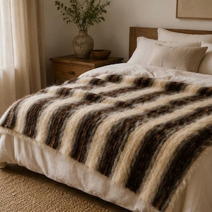 Peut inclure: Un plaid rectangulaire à la surface texturée et duveteuse, orné de rayures verticales alternées dans des tons crème, brun foncé et gris anthracite, posé sur un lit doté d'une parure blanche et de plusieurs oreillers aux tons neutres.