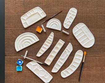 May include: A set of 10 white ceramic paint palettes in various shapes and sizes. The palettes have small indentations for holding paint. Two small paint pots are also included.