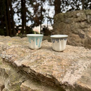Peut inclure: Deux petites tasses en céramique blanche avec un glaçage bleu et gris qui coule sur les côtés. Les tasses sont posées sur un rocher rugueux et texturé.