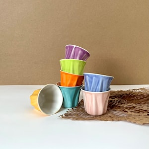 Puede incluir: Un juego de seis tazas de cerámica de colores apiladas en forma de pirámide. Las tazas son de diferentes colores, incluyendo morado, verde, naranja, azul y rosa. Las tazas están hechas de cerámica lisa y brillante y tienen un diseño único y ondulado.