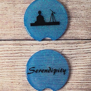 May include: Two blue glitter round coasters with black silhouettes. The top coaster features a person sitting with a telescope. The bottom coaster has the word "Serendipity" written on it.