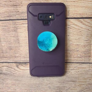 May include: A purple phone case with a textured design and a round, blue and green pop-up grip attached. The phone case has a camera and flash opening.