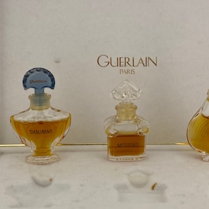May include: Three miniature perfume bottles in a white box. The bottles are labeled "Shalimar", "Mitsouko", and "Chamade". The box has the text "Guerlain Paris" printed on it.