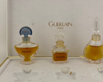 Può includere: Tre flaconi di profumo in miniatura in una scatola bianca. I flaconi sono etichettati "Shalimar", "Mitsouko" e "Chamade". La scatola ha la scritta "Guerlain Paris" stampata su di essa.