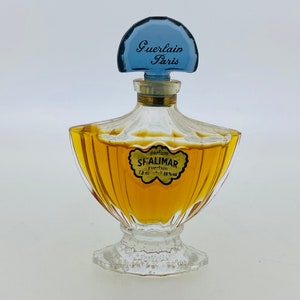 Può includere: Un flacone di profumo vintage Shalimar di Guerlain Paris. Il flacone è in vetro trasparente con un'etichetta dorata e un tappo in vetro blu. L'etichetta riporta "parfum Shalimar Guerlain 14 ml 3/8 fl oz."