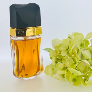 May include: A clear glass bottle of perfume with a gold cap and the word "Knowing" printed on the cap. The bottle is filled with a golden liquid and is sitting on a white surface. A bouquet of light green flowers is in the background.
