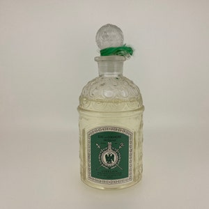 May include: A clear glass bottle of Guerlain Eau de Cologne Imperiale with a green and gold label. The bottle has a textured, ornate design and a clear glass stopper with a green ribbon.