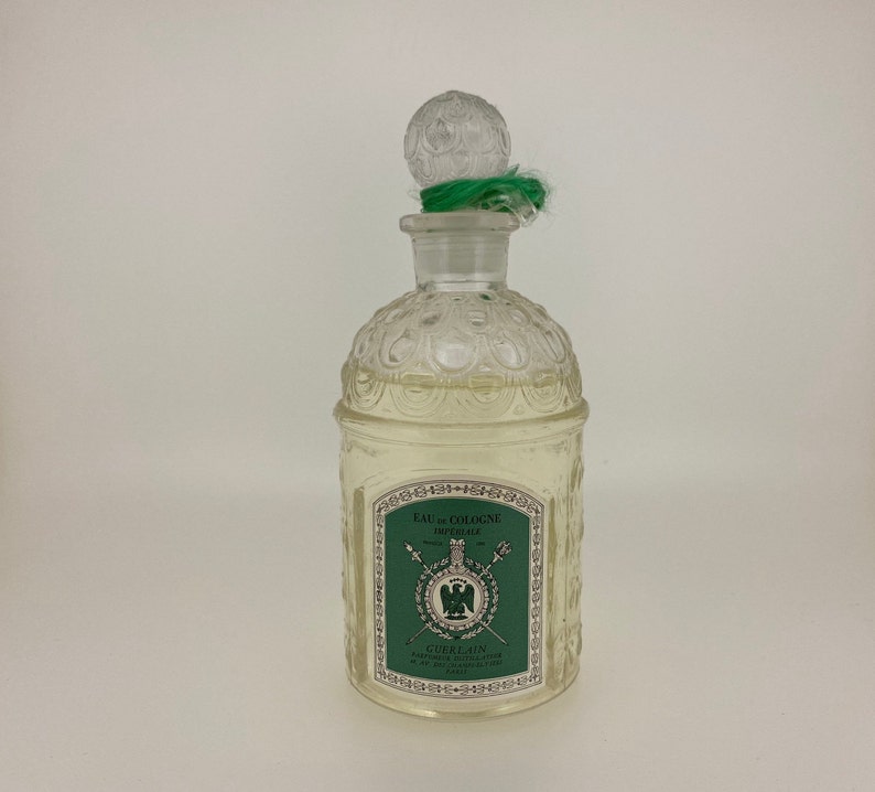 May include: A clear glass bottle of Guerlain Eau de Cologne Imperiale with a green and gold label. The bottle has a textured, ornate design and a clear glass stopper with a green ribbon.
