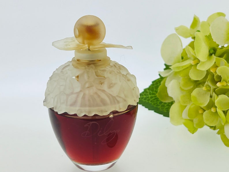 Może przedstawiać: Przezroczysta szklana butelka perfum z białą, kwiatową nakrętką i złotym wierzchołkiem. Butelka jest wypełniona ciemnoczerwonym płynem. Butelka stoi na białym tle.