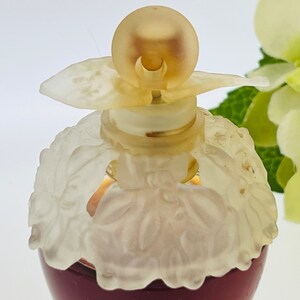 Może przedstawiać: Przezroczysta butelka szklana z białym wzorem kwiatowym i złotą zakrętką. Butelka jest wypełniona ciemnoczerwonym płynem.