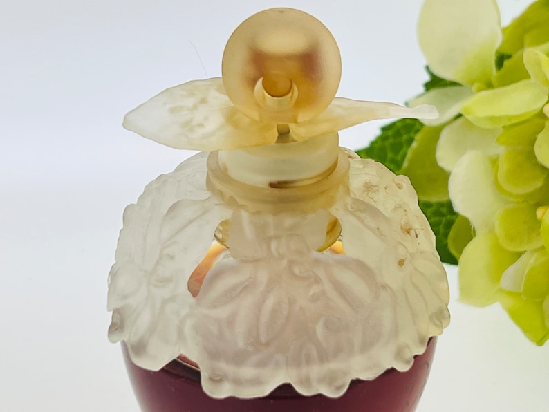 Może przedstawiać: Przezroczysta butelka szklana z białym wzorem kwiatowym i złotą zakrętką. Butelka jest wypełniona ciemnoczerwonym płynem.