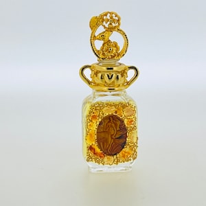 May include: A small, clear glass bottle with a gold-coloured top and a decorative design. The bottle is filled with a clear liquid and has a gold-coloured label with the word "IVI" on it.